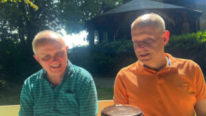 Zwei Männer unterhalten sich im Sonnenlicht. Im Hintergrund ist eine Hecke und ein Pavillion.