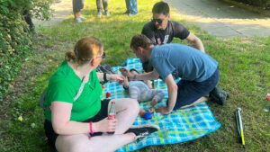 Eine Frau und zwei Männer sitzen auf einer Picknickdecke. Auf der Decke liegt ein Baby.
