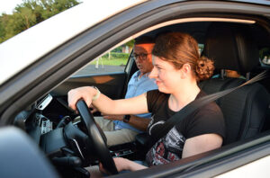 Eine blinde, junge Frau sitzt auf dem Fahrersitz eines Autos, neben ihr sitzt ein älterer Mann, beide sind angeschnallt.
