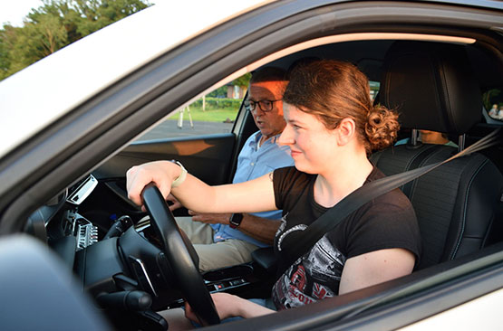 Eine blinde, junge Frau sitzt auf dem Fahrersitz eines Autos, neben ihr sitzt ein älterer Mann, beide sind angeschnallt.