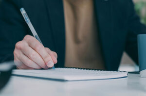 Eine Person in einem dunklen Blazer unterschreibt mit einem Stift ein Dokument. Sie sitzt an einem Schreibtisch, auf dem rechts eine blaue Tasse zu sehen ist.
