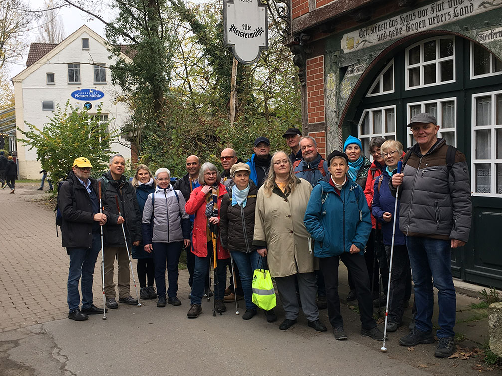 Eine Gruppe von 17 Menschen in Outdoor-Kleidung steht zusammen auf einem Gehweg in der Nähe eines historischen Backsteingebäudes mit einem Schild in altdeutscher Schrift darüber. Einige halten Blindenstöcke. Im Hintergrund sind Bäume und ein weiteres Gebäude zu sehen.