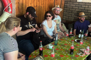 Fünf Personen sitzen um einen farbenfrohen Tisch mit leeren Tellern, Kaffeetassen und Colaflaschen. Die Gruppe scheint sich in einer ungezwungenen Umgebung zu unterhalten.