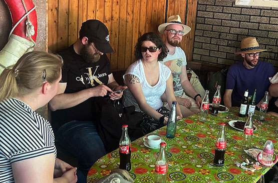 Fünf Personen sitzen um einen farbenfrohen Tisch mit leeren Tellern, Kaffeetassen und Colaflaschen. Die Gruppe scheint sich in einer ungezwungenen Umgebung zu unterhalten.