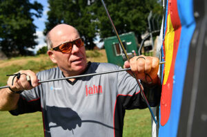 Ein Mann zieht im Freien Pfeile aus einer Bogenschießscheibe, er trägt ein graues Sporthemd und Fingerschutz an beiden Händen.