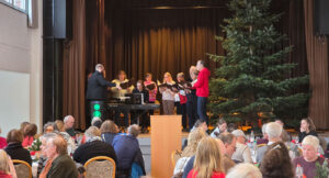 Ein Chor tritt auf der Bühne mit einem Pianisten und einem Dirigenten neben einem großen Weihnachtsbaum auf, während das Publikum an Tischen im Vordergrund sitzt.