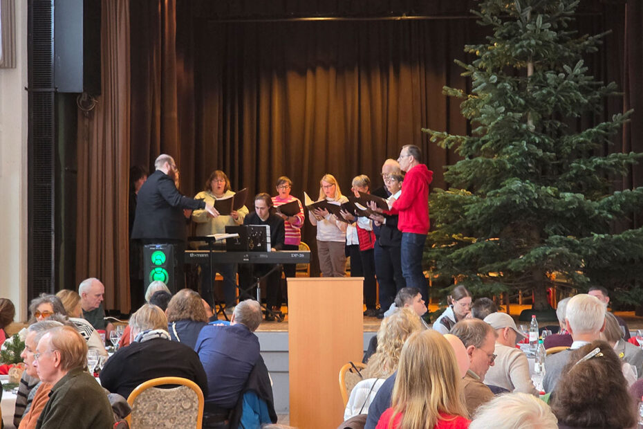 Ein Chor tritt auf der Bühne mit einem Pianisten und einem Dirigenten neben einem großen Weihnachtsbaum auf, während das Publikum an Tischen im Vordergrund sitzt.