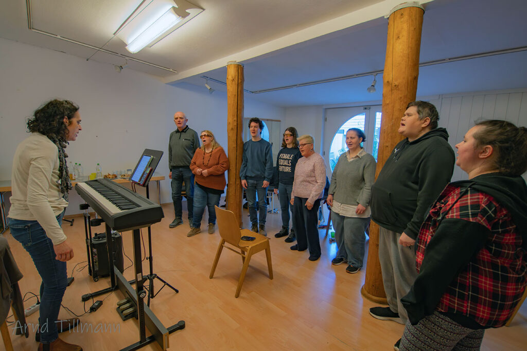 Eine Gruppe von Menschen steht in einem Halbkreis um ein Keyboard herum und blickt auf eine Frau, die sie in einem großen Raum mit Holzsäulen zu unterrichten scheint.