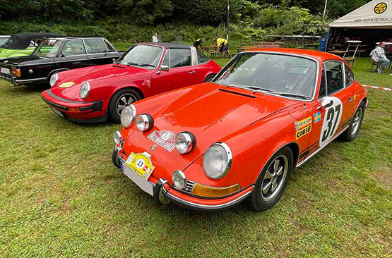 Drei Oldtimer sind bei einer Veranstaltung im Freien auf einer Wiese geparkt. Im Vordergrund steht ein roter Porsche 911 mit der Nummer 37. Im Hintergrund sind ein Zelt und Menschen zu sehen.