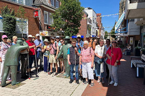 Eine Gruppe von Menschen mit Sehbehinderungen steht auf einer sonnigen Straße neben zwei Bronzestatuen, hält weiße Blindenstöcke in den Händen und trägt legere Sommerkleidung.