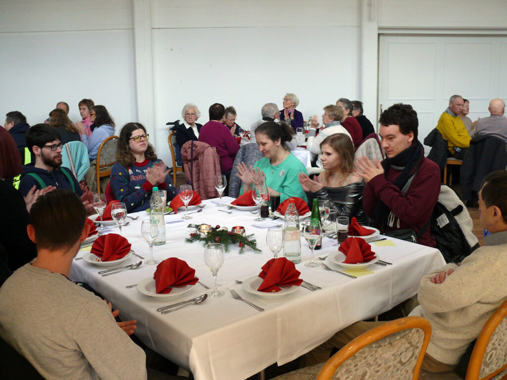 Vorne im Bild sitzen 8 Personen um einen gedeckten Tisch und applaudieren. Weitere Tische im Hintergrund sind besetzt.