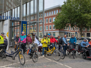 BSV- Eine Gruppe von Menschen mit Fahrrädern versammelt sich auf einem Gehweg in der Nähe eines Gebäudes mit Glaswänden und einem blau-gelben Schild. Im Hintergrund sind Bäume und geparkte Autos zu sehen.