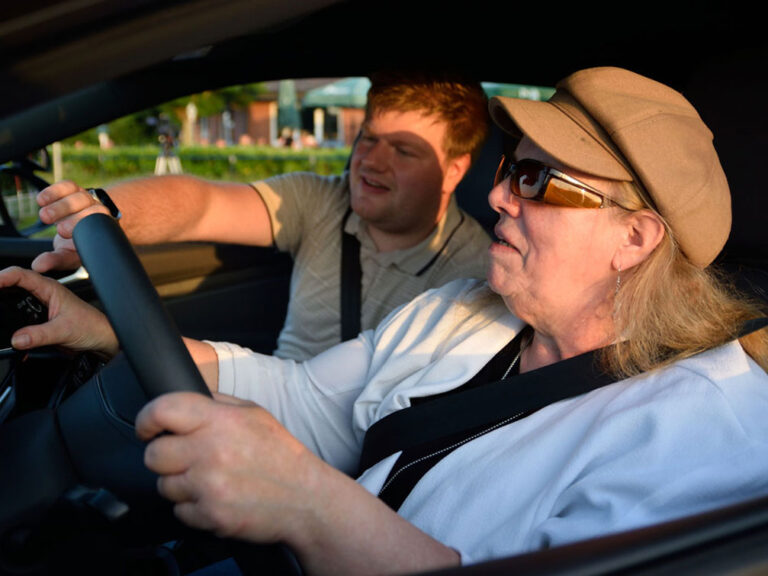 Eine ältere Frau mit Sonnenbrille und Hut sitzt auf dem Fahrersitz eines Autos und hält das Lenkrad fest, während ein Mann auf dem Beifahrersitz auf die Bedienelemente am Armaturenbrett zeigt.