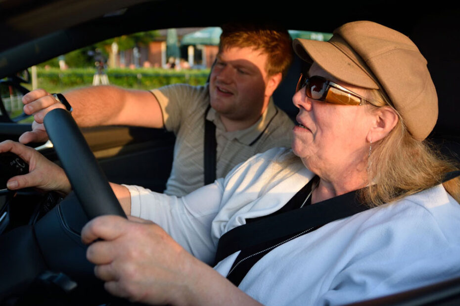 Eine ältere Frau mit Sonnenbrille und Hut sitzt auf dem Fahrersitz eines Autos und hält das Lenkrad fest, während ein Mann auf dem Beifahrersitz auf die Bedienelemente am Armaturenbrett zeigt.