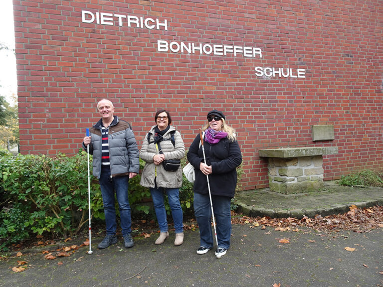 Zwei Frauen und ein Mann stehen nebeneinander vor einer Klinkerwand mit dem Schriftzug Dietrich Bonhoeffer Schule.