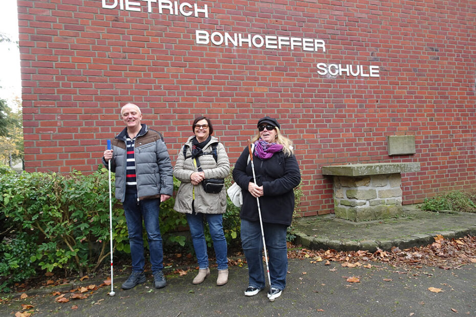 Zwei Frauen und ein Mann stehen nebeneinander vor einer Klinkerwand mit dem Schriftzug Dietrich Bonhoeffer Schule.