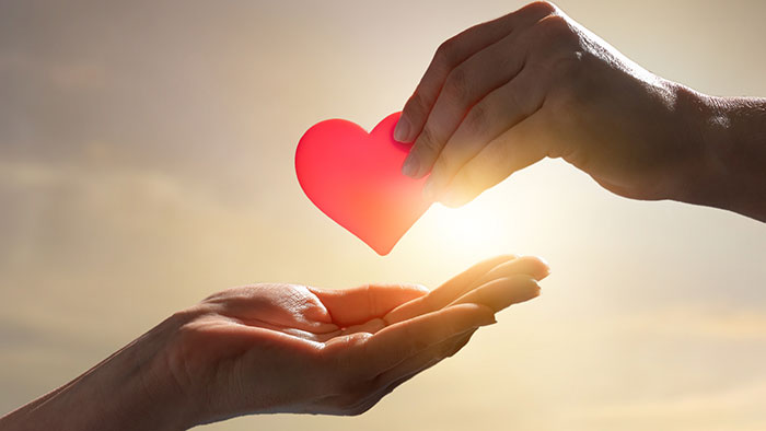 Vor einem hellen, sonnenbeschienenen Hintergrund übergibt eine Hand einen roten, herzförmigen Gegenstand an eine andere Hand.