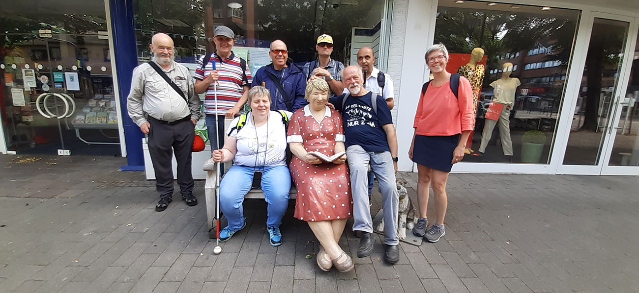 Eine Gruppe von acht Erwachsenen posiert im Freien um eine sitzende Statue einer Frau, die ein Buch hält, vor einem Schaufenster mit Schaufensterpuppen.