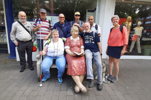 Eine Gruppe von acht Personen posiert im Freien um eine sitzende Statue einer Frau, die ein Buch liest, wobei im Hintergrund ein Schaufenster und Schaufensterpuppen zu sehen sind.