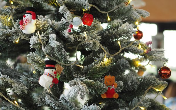 Der Ausschnitt eines Weihnachtbaums in der Nahaufnahme. Seine Zweige sind mit Schnee bedeckt. Rote Weihnachtskugeln, ein Teddy und kleine Schneemänner mit schwarzen Zylinderhüten zieren siene Zweige.