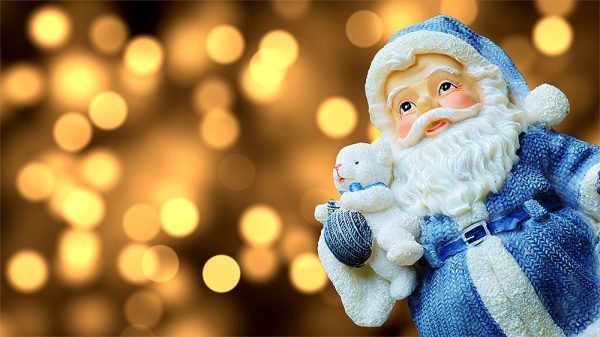 Im rechten Teil des Bildes befindet sich ein Weihnachtsmann in der Nahaufnahme. Er hält einen weißen Teddybären im rechten Arm. Das besondere ist, Santa Claus trägt einen blauen Mantel und eine blaue Mütze, heute mal nicht in rot. Im Hintergrund sind schemenhafte goldene Lichter zu sehen. Sind es vielleicht Kerzen oder Christbaumkugeln?