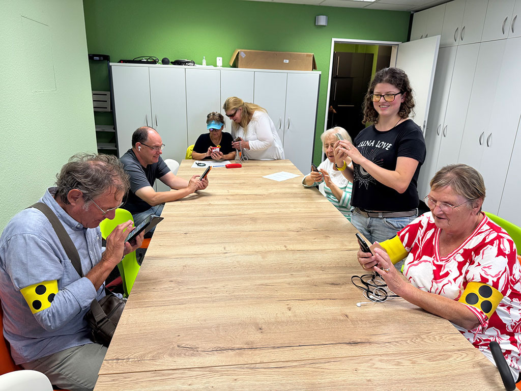 Eine Gruppe älterer Erwachsener sitzt während einer iPhone-Schulung an einem Tisch und benutzt Smartphones, wobei zwei Ausbilder assistieren. Mehrere Teilnehmer tragen gelbe Armbinden mit schwarzen Punkten. Der Raum hat grüne Wände und ist mit weißen Schränken ausgestattet.