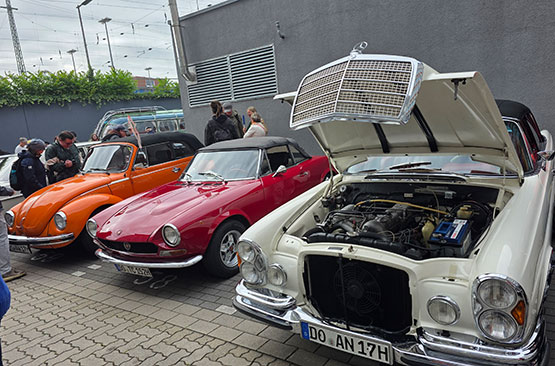 Bei einer Veranstaltung im Freien stehen drei Oldtimer nebeneinander; bei dem Auto auf der rechten Seite ist die Motorhaube geöffnet, sodass der Motor zu sehen ist. Um die Fahrzeuge herum haben sich Menschen versammelt, die sie sich ansehen.