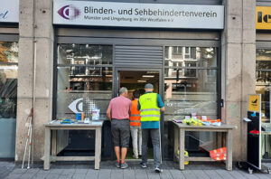 Zwei Männer betreten die Beratungsstelle des Blinden- und Sehbehindertenvereins Münster und Umgebung. Links und rechts neben der Eingangstür steht jeweils ein Tisch mit Informationsmaterial.